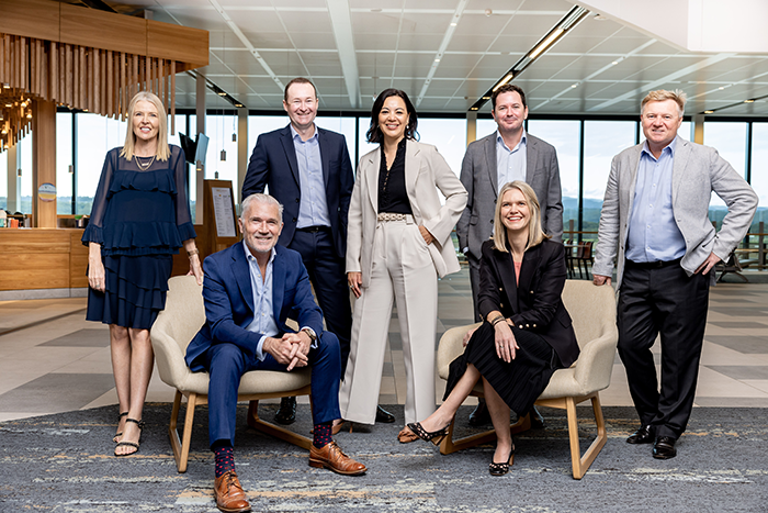 Seven professionally dressed individuals pose for a group photo in a modern airport lounge or office setting, with large windows and natural light in the background.