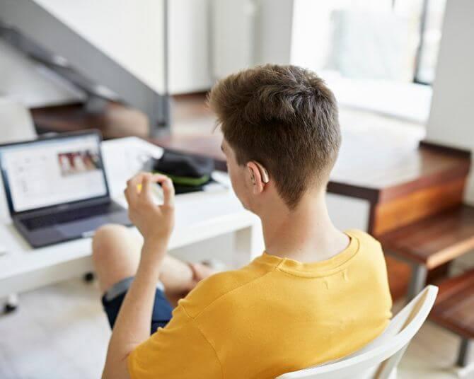 A young man wearing a yellow T-shirt sits at a white desk in a modern home interior, looking at a laptop screen.