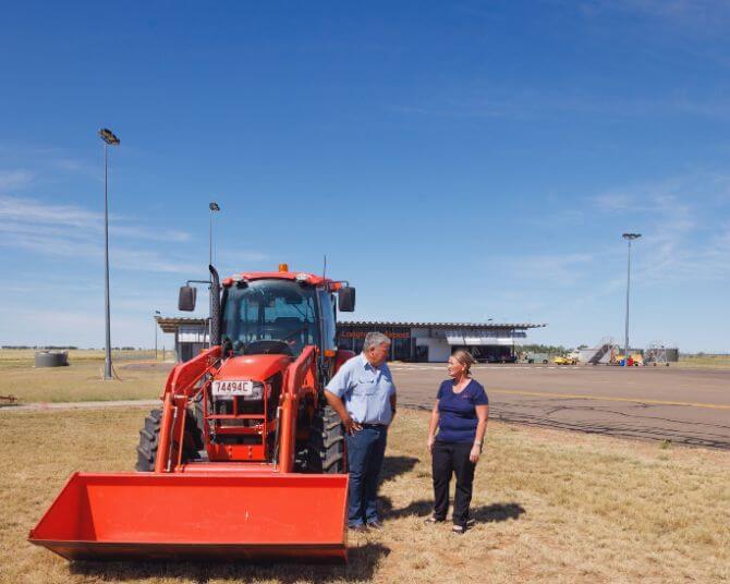 Airport team members standing beside a bright orange tractor at Longreach Airport, with terminal building and airfield in the background under clear blue sky.