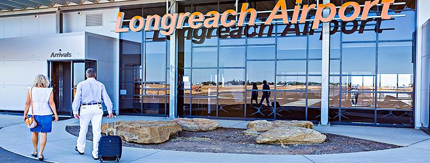 Two travelers walking toward the Arrivals entrance at Longreach Airport, with large glass windows reflecting the runway and orange Longreach Airport signage above.