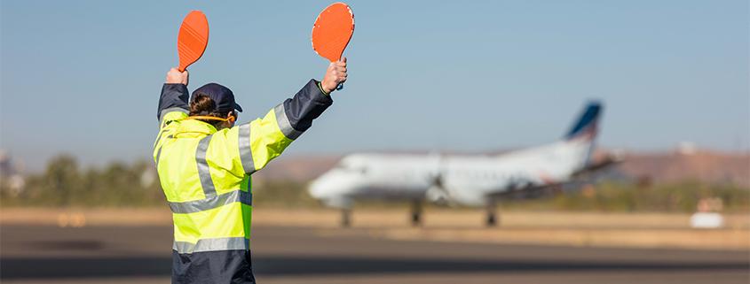 Ground crew member in high-visibility jacket guiding an aircraft on the runway using orange marshalling paddles at Longreach Airport.
