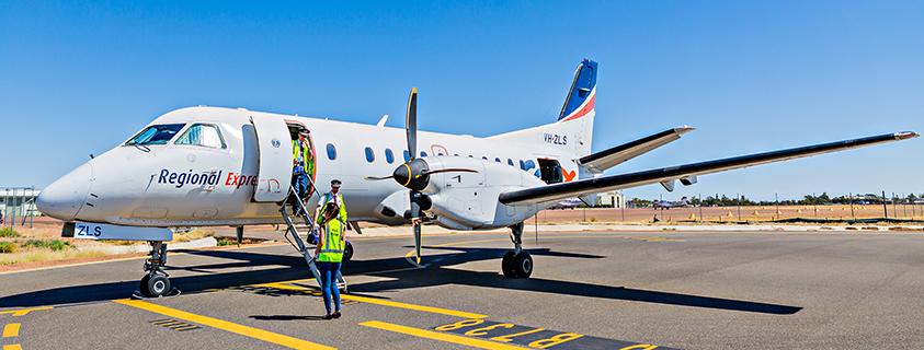 Regional Express aircraft parked on the tarmac at Longreach Airport with ground crew assisting passengers boarding via the aircraft stairs.