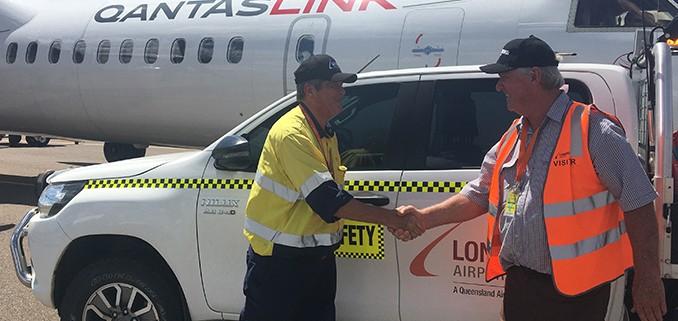 Two people shaking hands beside a Longreach Airport safety vehicle, with a QantasLink aircraft in the background.