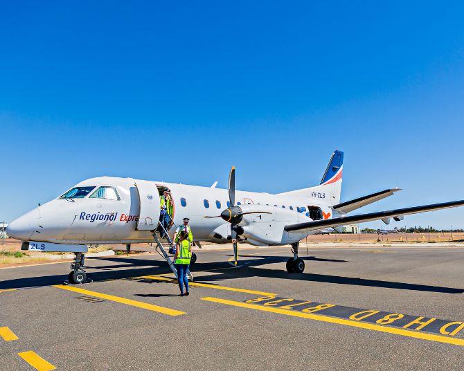A Regional Express aircraft parked on the tarmac under a clear blue sky, with passengers boarding via the front stairs and ground crew standing nearby.