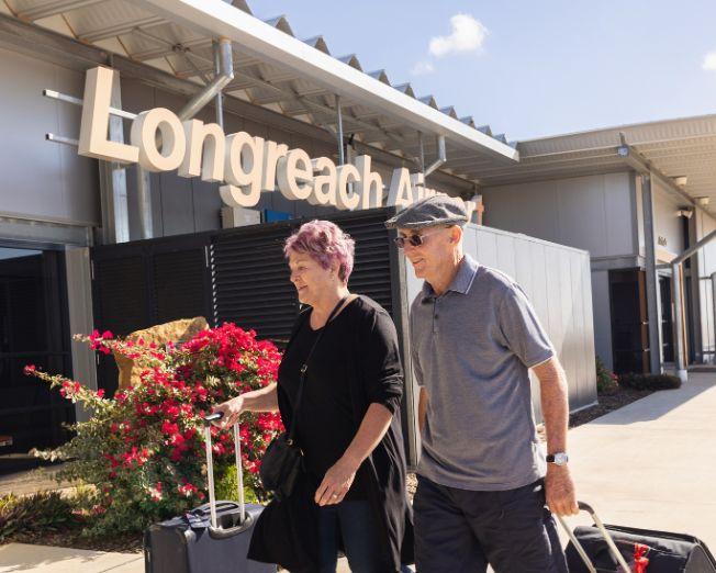Two travelers with luggage walking outside Longreach Airport terminal under a clear sky.