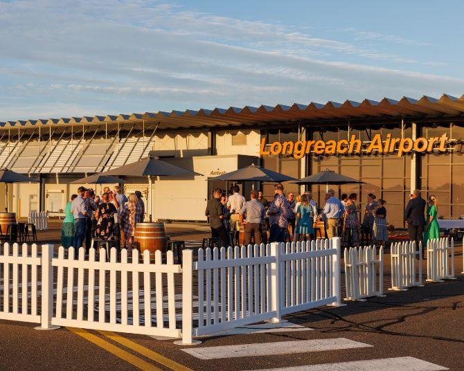 Community event outside Longreach Airport terminal with people gathered near tables and umbrellas, enclosed by a white picket fence.