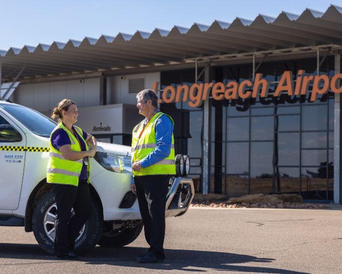 Two airport staff wearing high-visibility vests standing beside a white airport vehicle in front of the Longreach Airport terminal building.