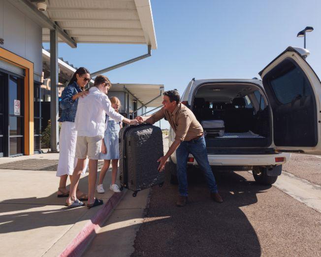 Person handing a large suitcase to a traveler at the passenger drop-off area outside Longreach Airport terminal, with an open SUV parked at the curb.