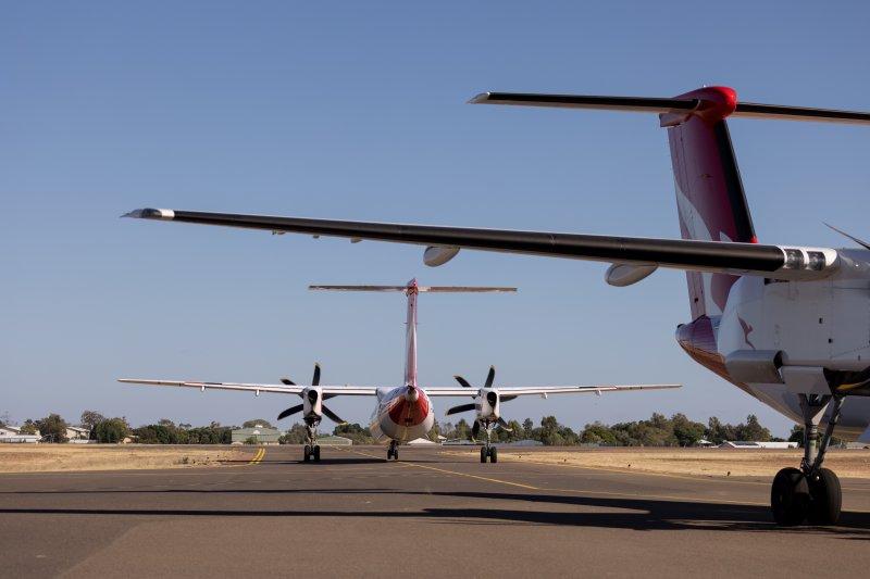 Two QantasLink aircraft on the taxiway at Longreach Airport, viewed from behind with wings and tail visible under a clear blue sky.