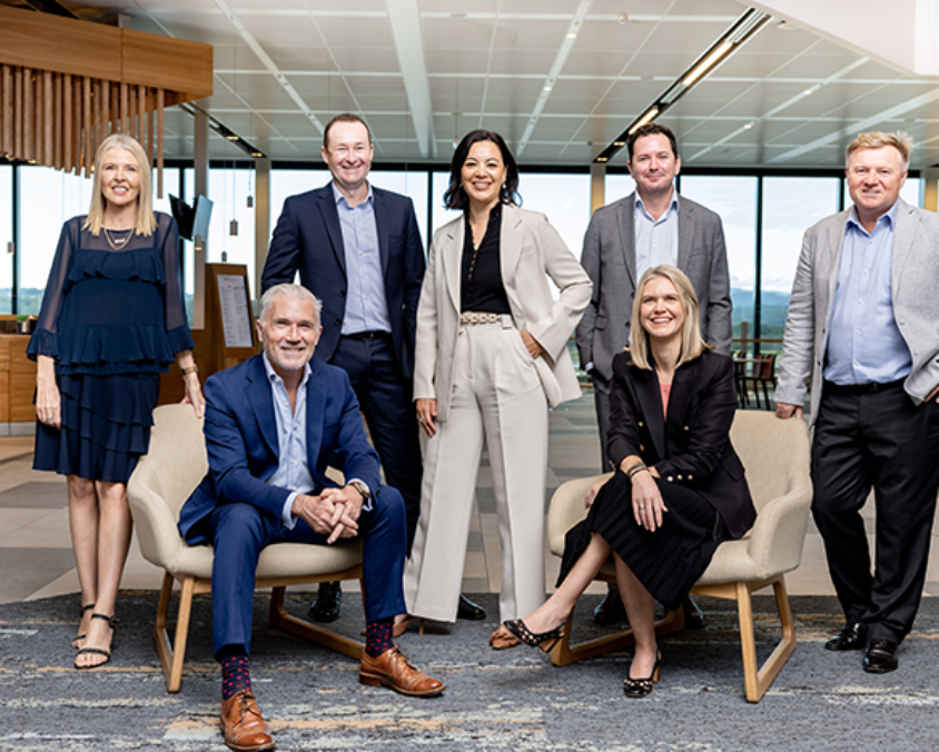 Seven professionally dressed individuals pose for a group photo in a modern airport lounge or office setting, with large windows and natural light in the background.