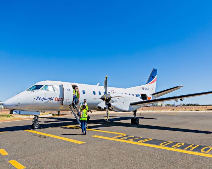 A Regional Express aircraft parked on the tarmac under a clear blue sky, with passengers boarding via the front stairs and ground crew standing nearby.
