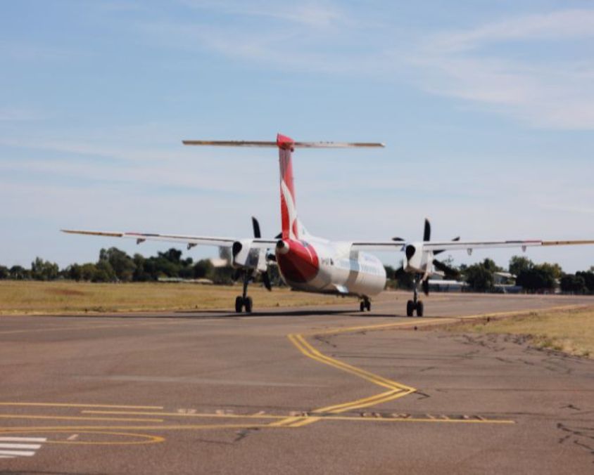 A QantasLink turboprop aircraft taxiing along the runway, viewed from behind with its tail and wings visible against a light blue sky.