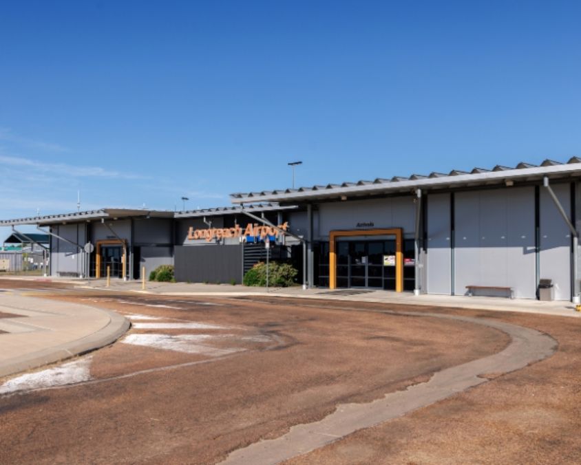 Exterior view of Longreach Airport terminal with arrivals entrance and curved roadway in front under a clear blue sky – click to explore the 360° VR walkthrough by Queensland Airports Limited