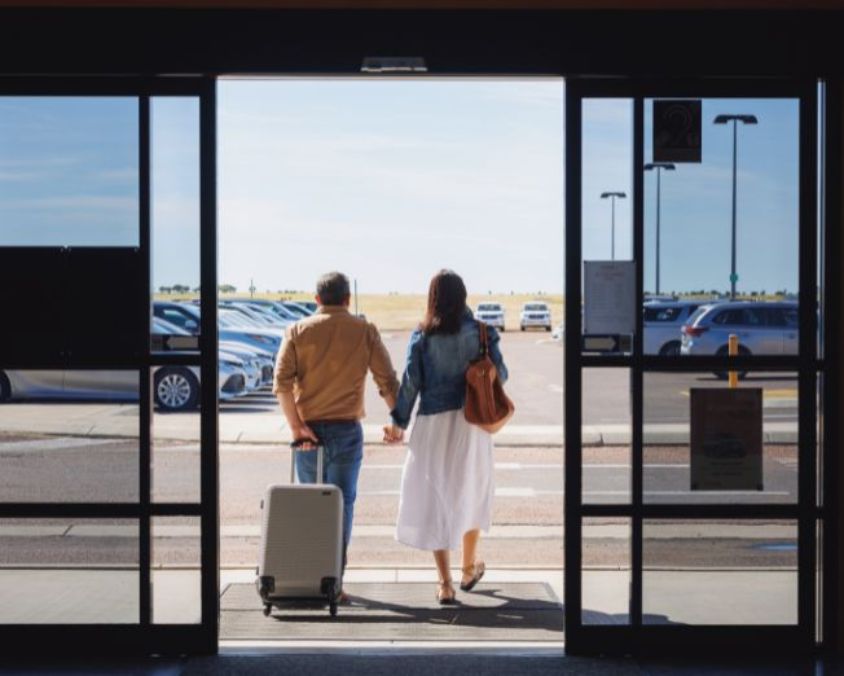 Two travelers holding hands and walking out of Longreach Airport terminal toward the car park, with a suitcase and clear blue sky in the background