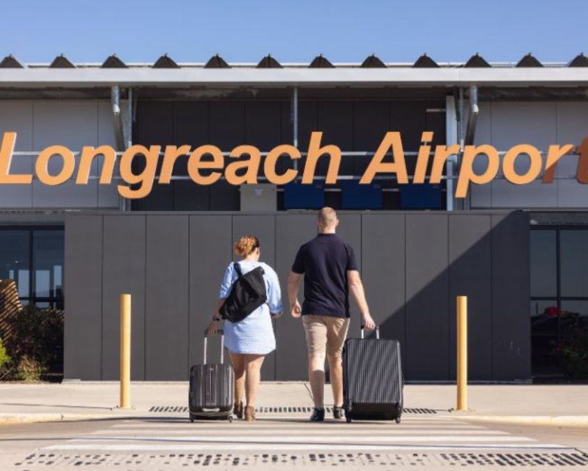 Two travelers with suitcases walking toward the entrance of Longreach Airport terminal under clear blue sky.