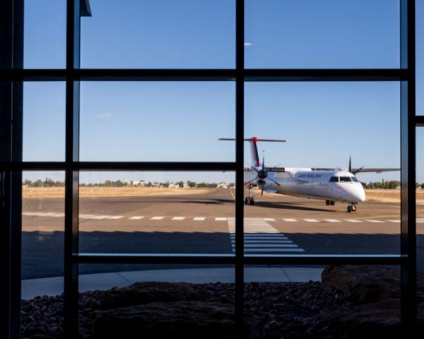 QantasLink aircraft taxiing on the runway at Longreach Airport in Queensland, viewed through terminal windows under a clear blue sky