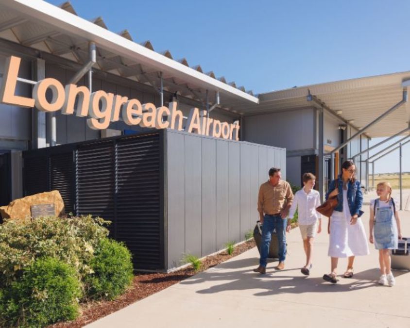 Family walking outside Longreach Airport terminal building under clear blue sky.