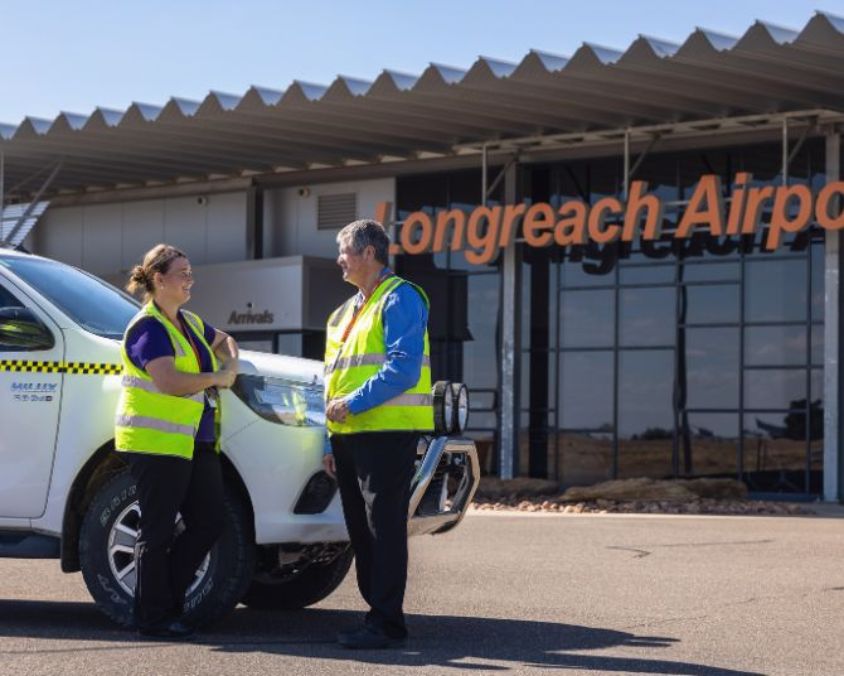 Two airport staff wearing high-visibility vests standing beside a white airport vehicle in front of the Longreach Airport terminal building.