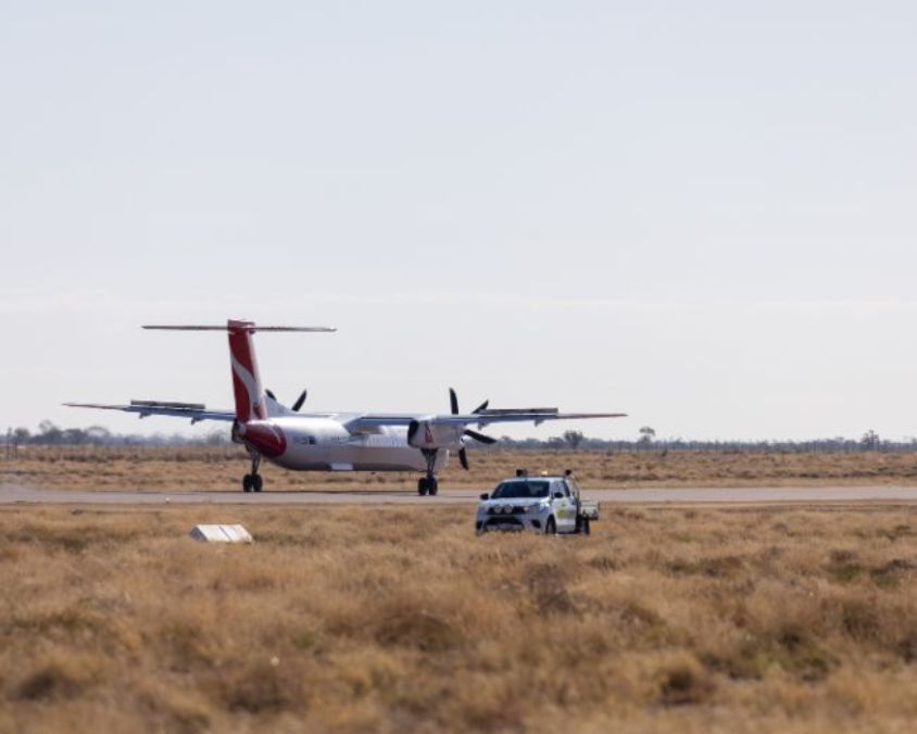 A QantasLink aircraft taxis along the runway at Longreach Airport while an airside safety vehicle is parked nearby on the airfield.