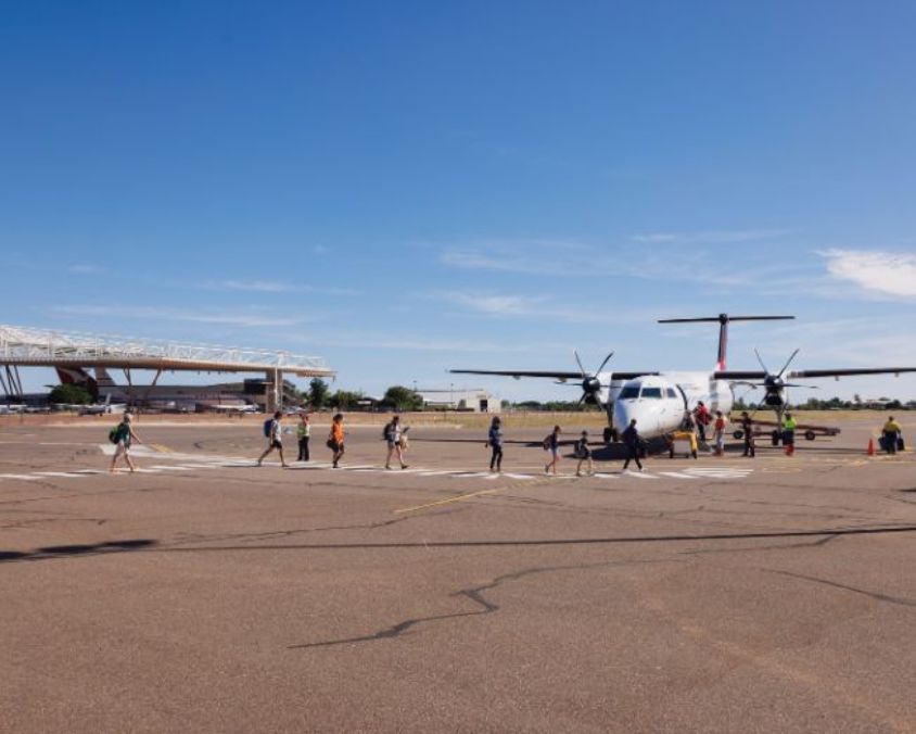 A line of passengers walks along a designated path on the tarmac toward a white turboprop aircraft at Longreach Airport, with ground crew guiding them and the terminal visible in the distance.