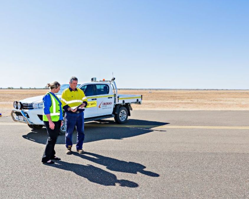 Airport safety staff stand on the Longreach Airport runway discussing paperwork beside a safety vehicle, with clear skies and outback landscape in the background.