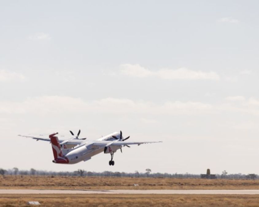 A QantasLink aircraft takes off from Longreach Airport, rising over the flat outback terrain under a pale sky.