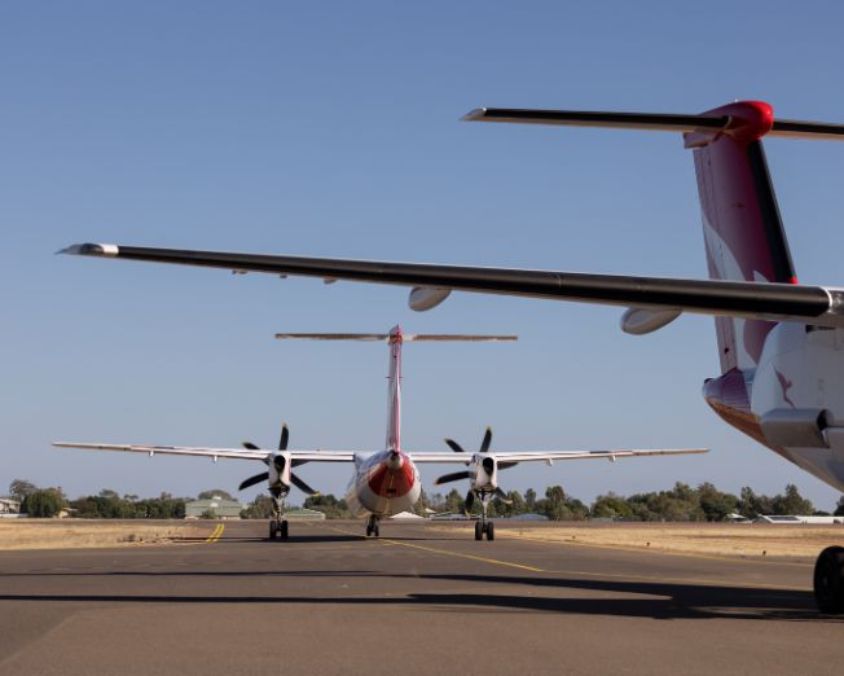Two QantasLink aircraft on the taxiway at Longreach Airport, viewed from behind with wings and tail visible under a clear blue sky.