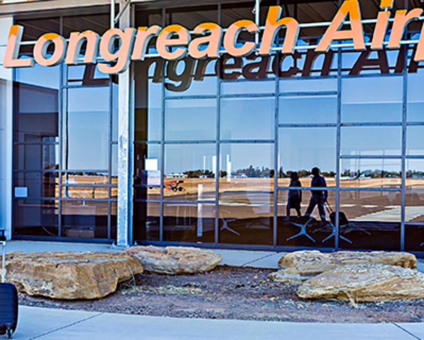 Two travelers walking toward the Arrivals entrance at Longreach Airport, with large glass windows reflecting the runway and orange Longreach Airport signage above.