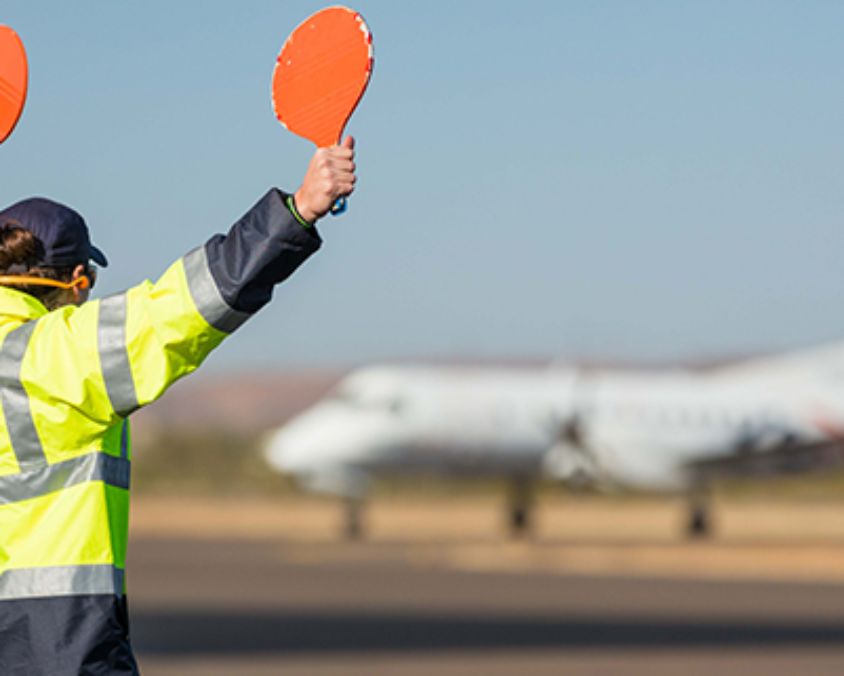 Ground crew member in high-visibility jacket guiding an aircraft on the runway using orange marshalling paddles at Longreach Airport.