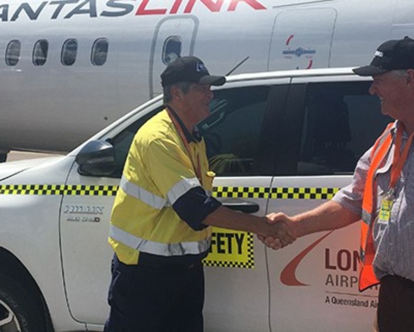 Two people shaking hands beside a Longreach Airport safety vehicle, with a QantasLink aircraft in the background.